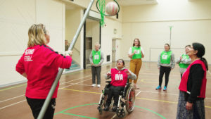 Wheelchair netball player shoots a ball through a hoop being lowered by a coach.