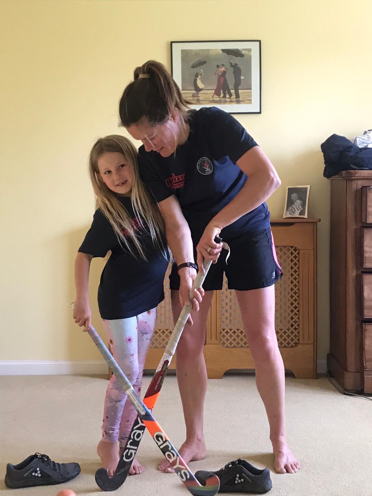 A mother and daughter standing in a living room holding hockey sticks diagonally across each other to form a X shape.