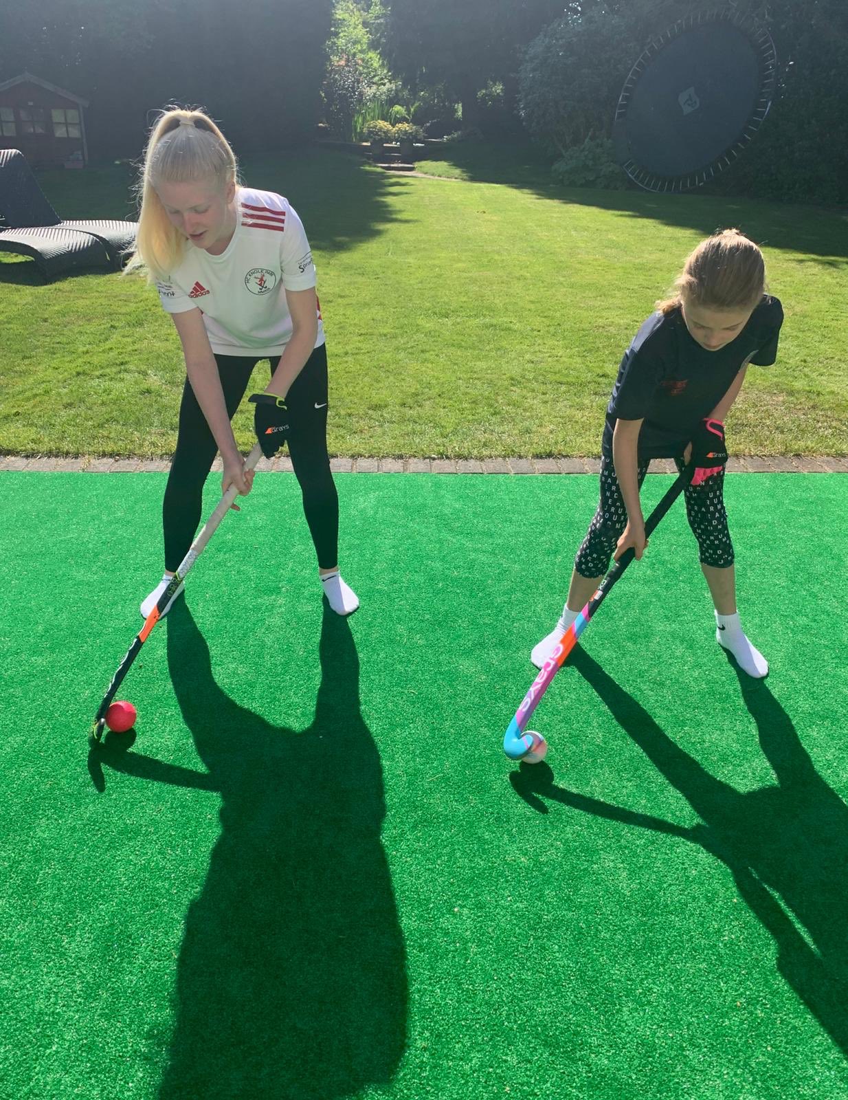 Two girls standing on grass in a garden using hockey sticks to move a hockey ball.