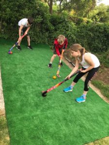 A girl and two boys standing on artificial grass in a garden using hockey sticks to move hockey balls.