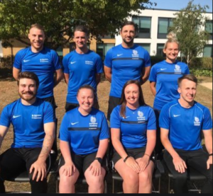 The Brompton Academy PE department on a grass field outside their school. Eight people wearing blue branded T shirts, four sitting on chairs with four standing behind them.