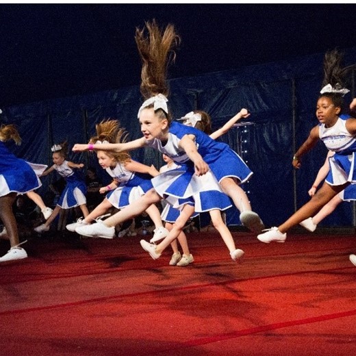 Group of girls in cheerleader outfits jumping in synchronised star jump poses.