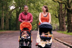 two women walking and smiling and pushing their babies in prams in a park