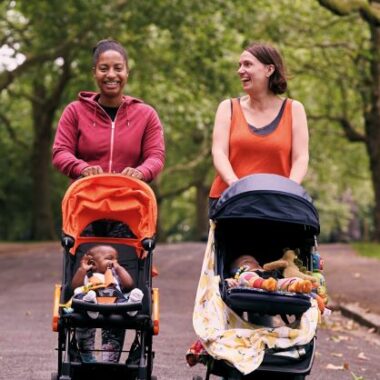 two women walking and smiling and pushing their babies in prams in a park