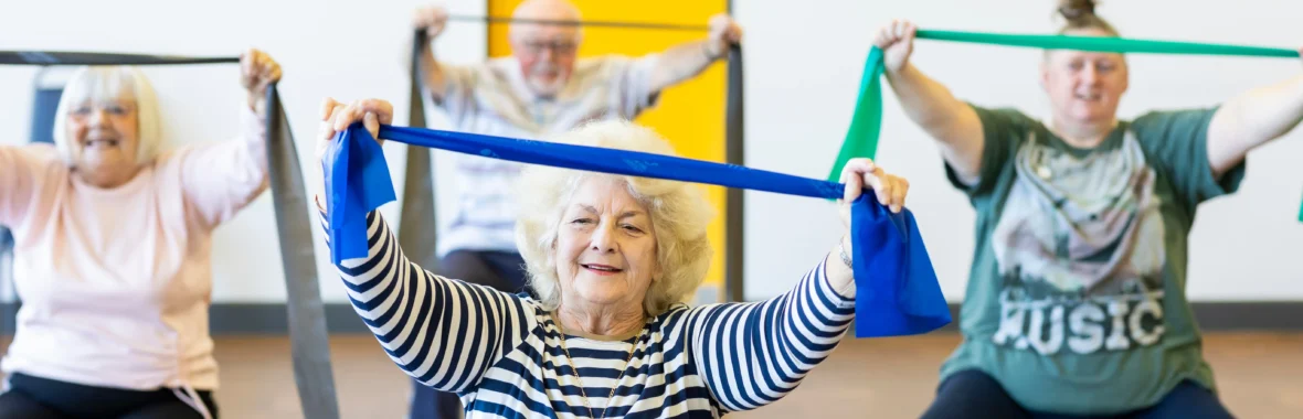 Women in an exercise class sat on chairs holding up resistance bands