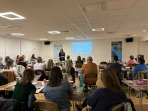 group of about 40 people sat in a room listening to a person standing at the front giving a presentation